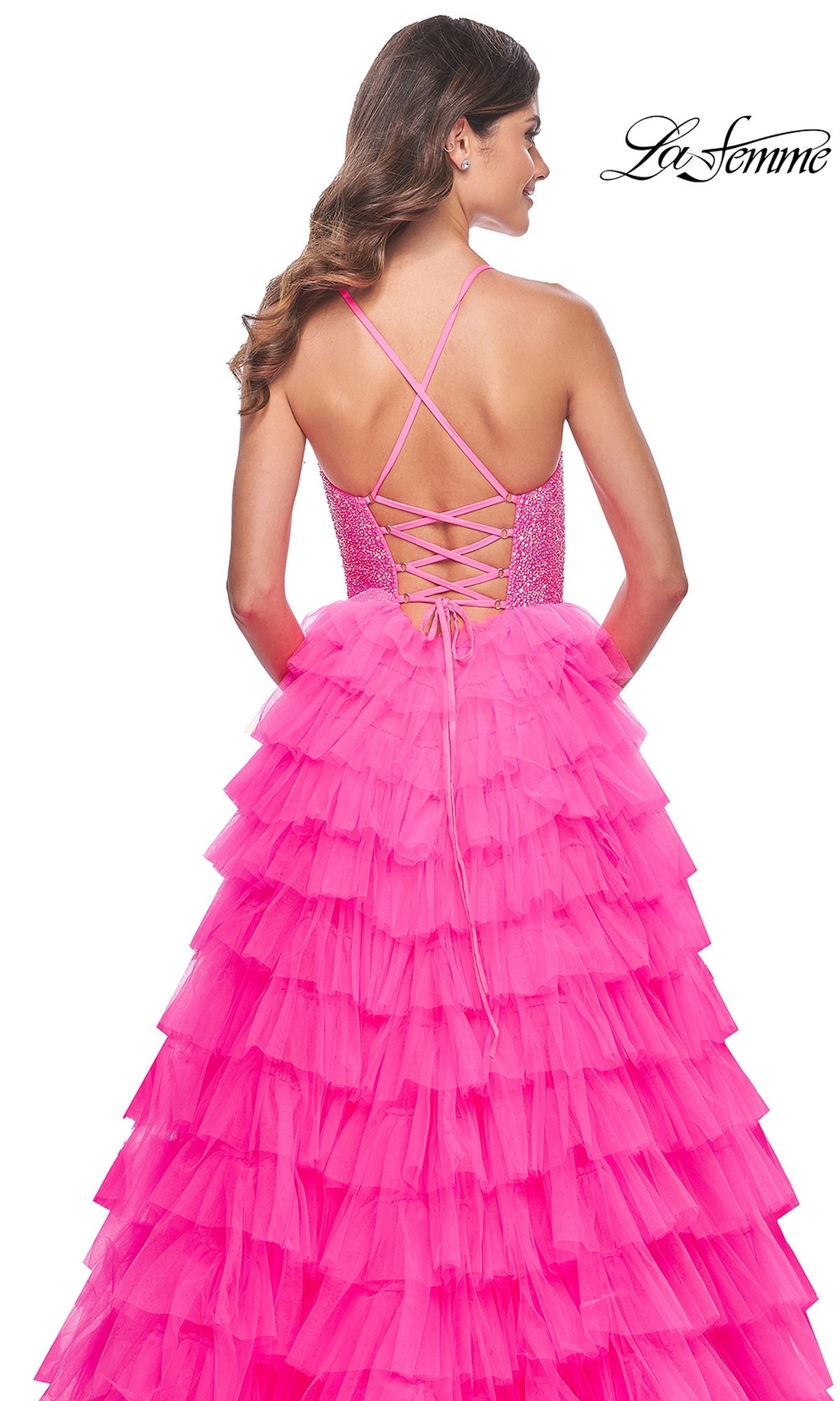 Woman in the La Femme Long Tiered A-Line Prom Dress 32335, featuring bright pink ruffles, a lace-up back, crisscross straps, and a thigh-high side slit, stands facing away from the camera.